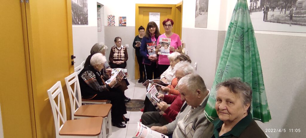 na korytarzu siedzą  seniorzy. czytają Głos Seniora. przy seniorach stoją dwie ambasadorki Głosu Seniora i trzymają gazety.