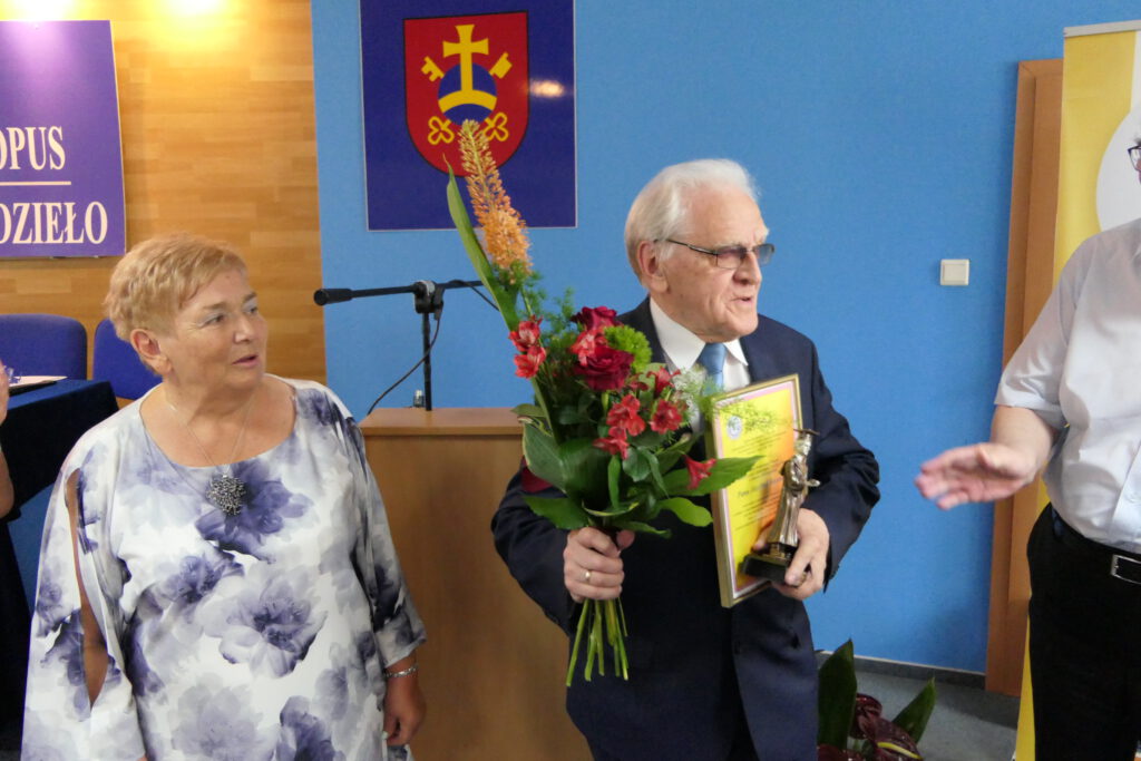 Józef Kozan otrzymuje godność honorowego Prezesa Stowarzyszenia OUTW. Trzyma kwiaty, statuetkę i dyplom. Obok niego stoją dwie osoby.